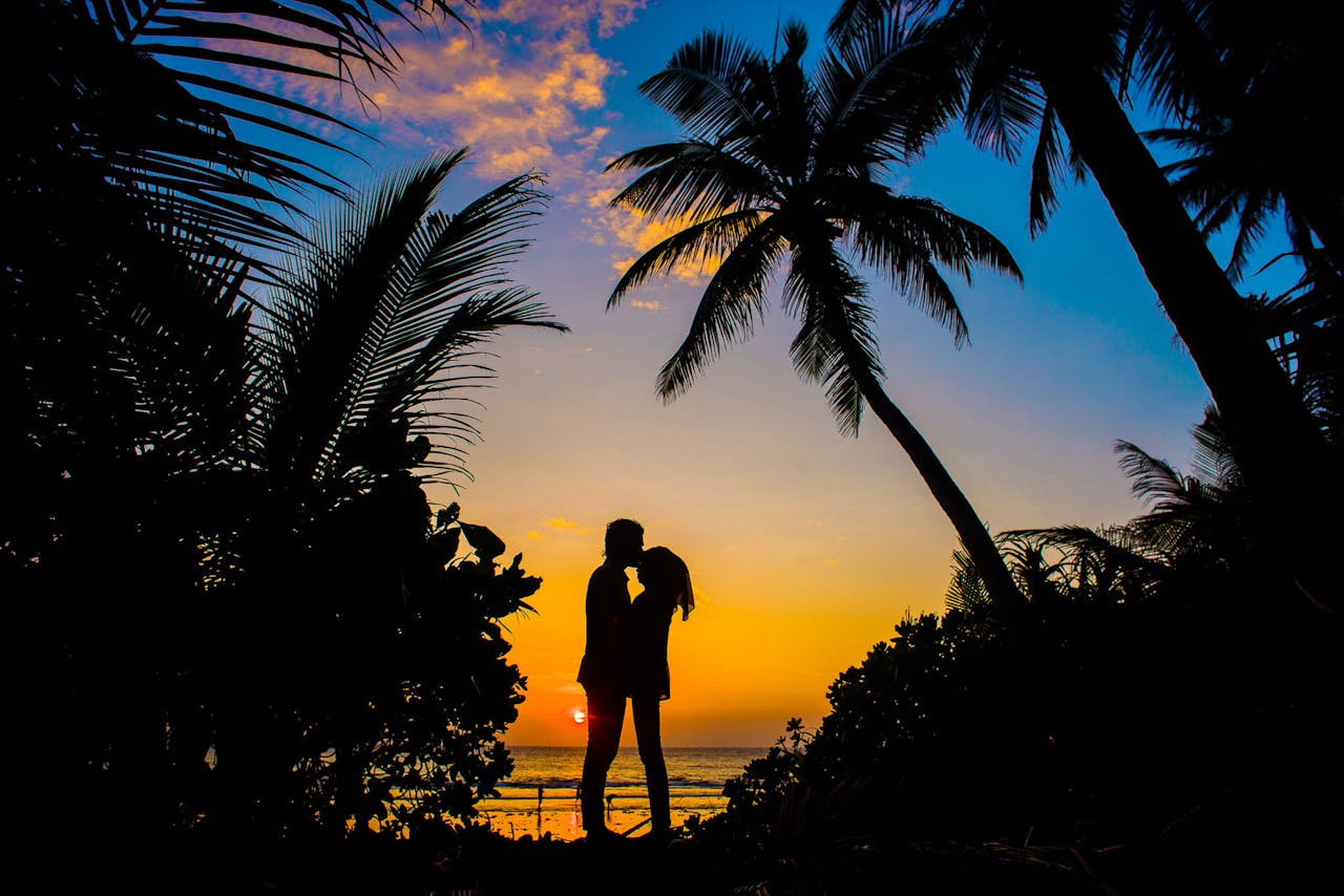 who-we-are Silhouette of a couple sharing a romantic moment on a tropical beach at sunset with palm trees.