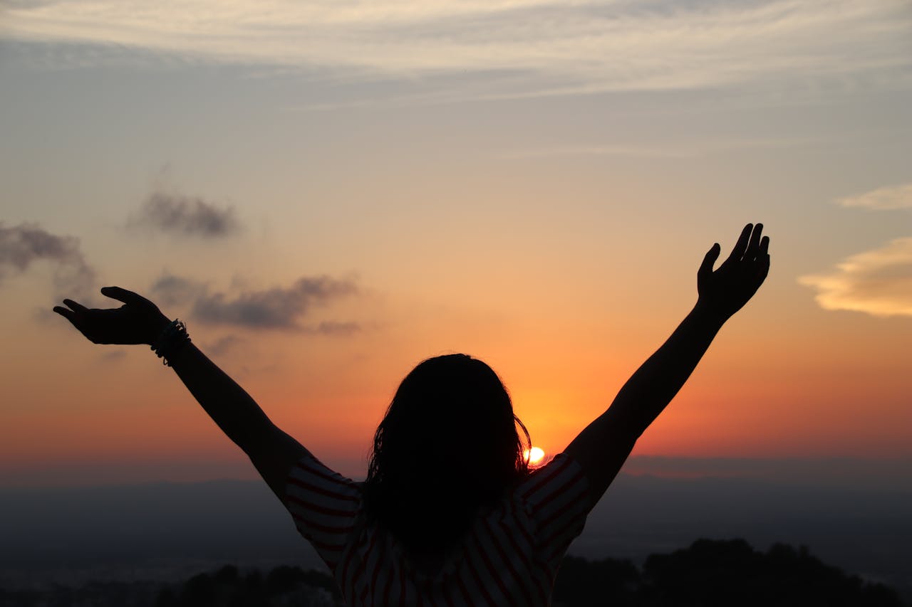 heros-img Silhouette of a woman with raised arms embracing the sunset scenery from a hilltop view.
